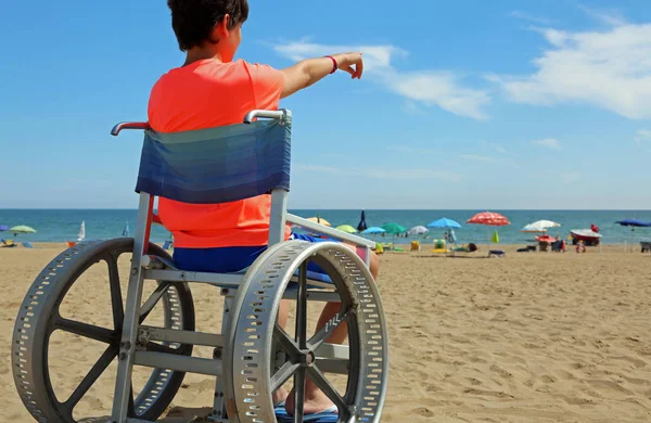 boy on a special wheelchair indicates the horizon of the sea on the sandy beach in summer