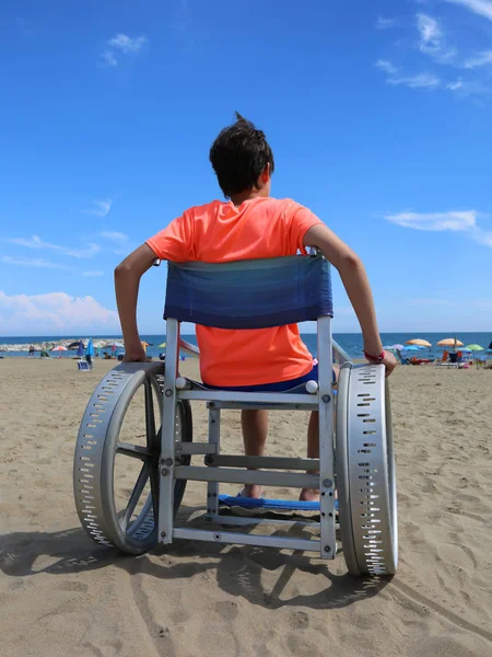 Tekerlekli sandalye, tatil köyünün plajında genç bir çocukla. — Stok fotoğraf