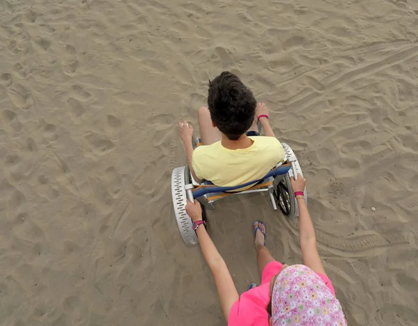 Klein meisje beweegt de rolstoel op het strand met een jonge jongen — Stockfoto
