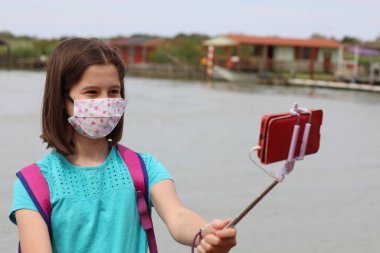 Little girl with surgical mask because of the Corona Virus takes a selfie with the smart phone and the selfiestick clipart