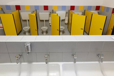Anaokulunda dört musluğu olan uzun bir lavabo. Tuvaletleri ve sarı kapısı olan çocuksuz bir çocuk.
