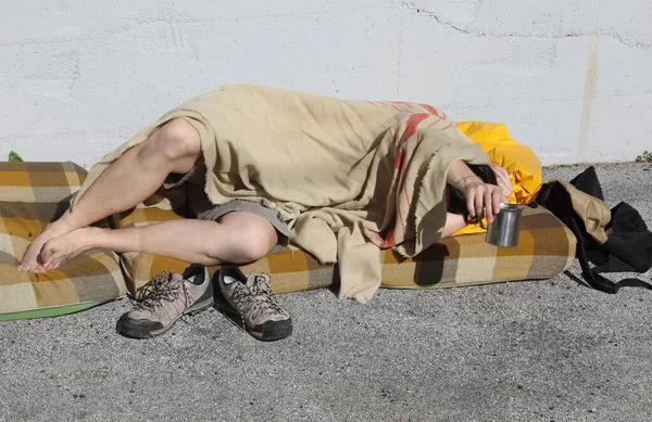 homeless man begging passersby in a city street while resting under a dirty blanket and a dirty mattress