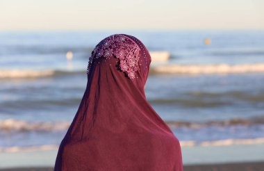 Başında mor peçeli genç bir kadın ailesinin denizden gelişini bekliyor.