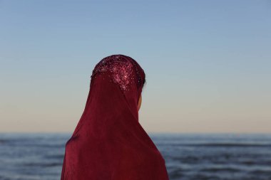 Kafasında deniz kenarında mor peçeli genç bir Arap kadın teknenin gelişini bekliyor.