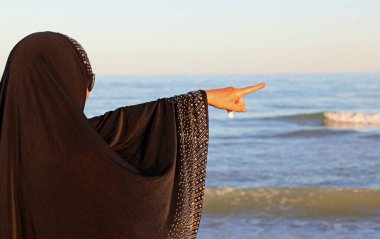 Kafasında siyah peçeyle genç bir kadın ailesinin denizden gelişini bekliyor ve işaret parmağıyla ufku işaret ediyor.