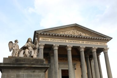 Bulutlu bir gökyüzünün altında, elinde Kutsal Kase olan bir kadının heykeliyle birlikte büyük sütunları ve heykelleri olan tarihi neoklasik bir bina.