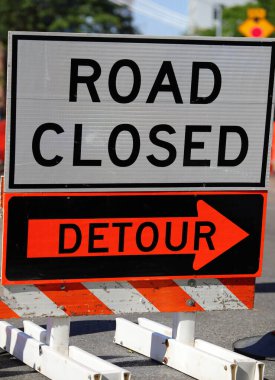 Büyük yol kapalı tabelası, tali bir ok ve turuncu inşaat varilleri bir şehir caddesinde.