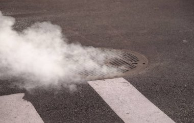 İşlek bir caddede dökme demir rögar kapağından beyaz duman çıkıyor.