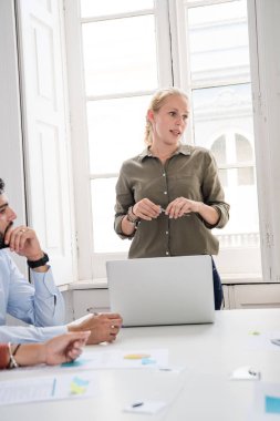 Yeşil gömlekli sarışın saçlı kadın kalem tutan biri onu tarafına konuşurken bir ofiste duruyor