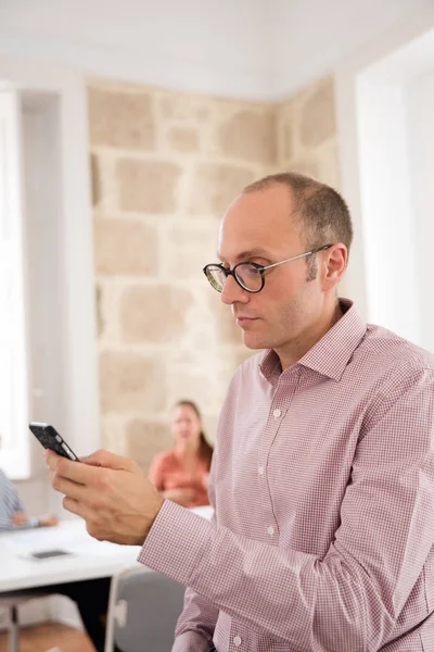 Interested man in a pink shirt is holding a black cellphone and leaning against a desk sending a text message