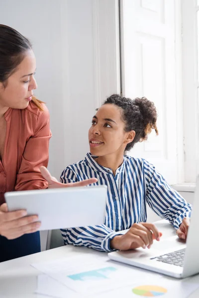 Two women working in an office, one sitting and the other is standing next to her and they are talking