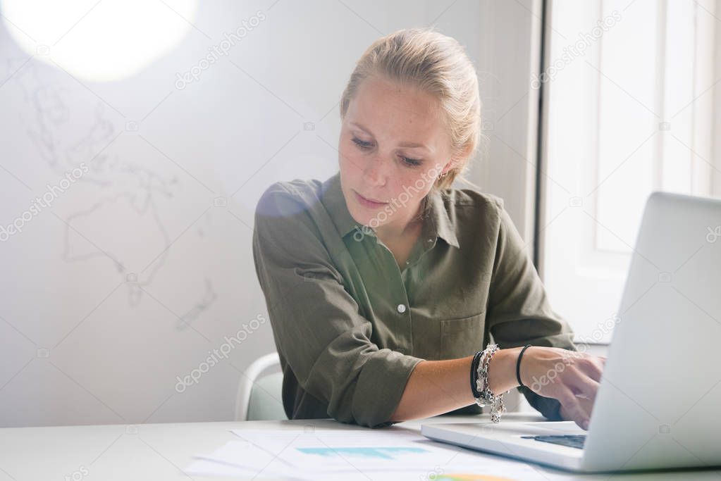 Blonde woman typing on a laptop as she looks at the document next to her, she is in an office