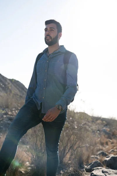 Man standing on a path with tall grass surrounded by a low hill and rocks looking at the horizon