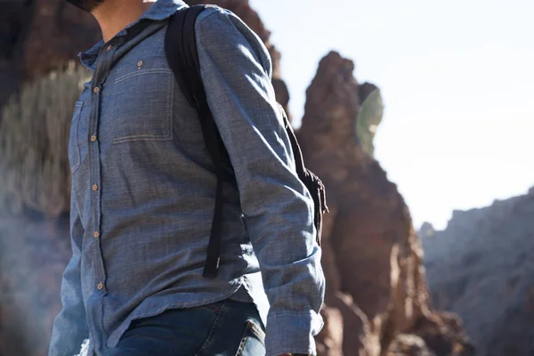 Side view of a man in a blue shirt walking past an outcrop of rocks on a mountain, carrying a backpack