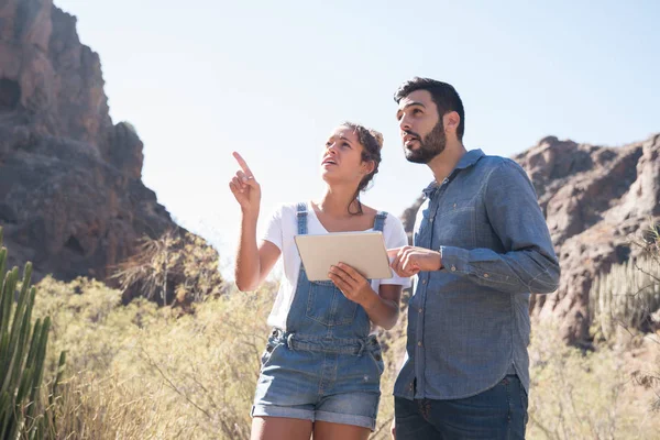 Couple standing on a mountain path next to cactus, they are looking up and pointing at the sky