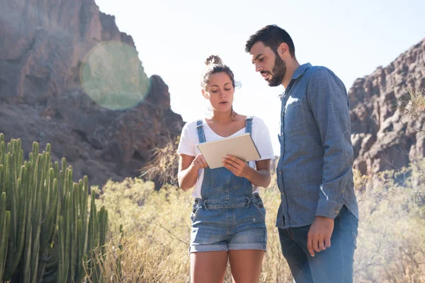 Man and woman standing on a mountain path next to long brown grass and cactus as the woman holds a pc tablet