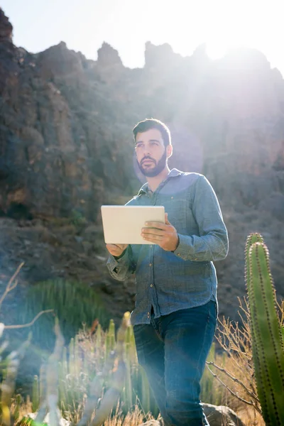 Man standing on a mountain path next to a cactus, holding a pc tablet and looking ahead