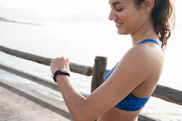 Woman smiling as she look at her watch as she stands by a wooden fence at the beach