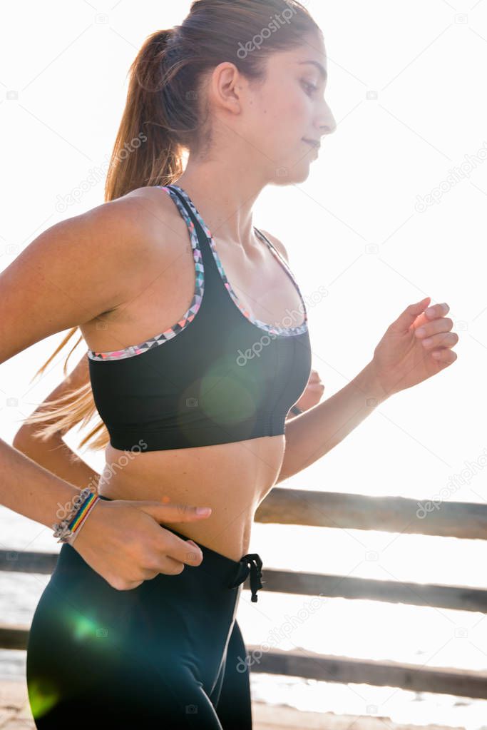Young woman running on the beach front, next to a wooden fence, with her pony tail flowing in the breeze