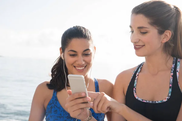 Two women standing by the ocean one has ear phones in her ears and the other is pointing at a cell phone