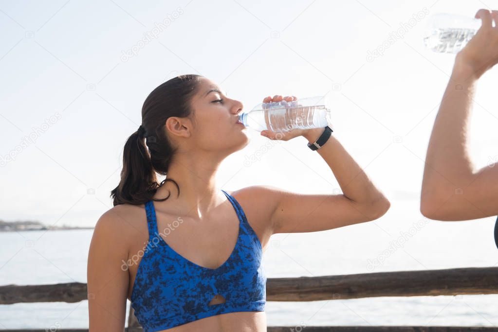 Two women are standing next to wooden fence overlooking the ocean and drinking water from plastic bottles