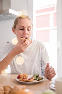 Bir mutfak bir masada oturur gibi kadın bir kruvasan yiyor, o yemeğini kahvaltıda aşağı bakıyor