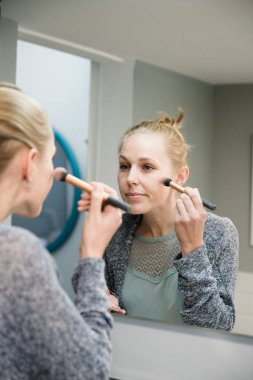 Gri bir forma giyen ve onun yüzüne makyaj uygulamak bir ayna önünde duran genç sarışın kadın
