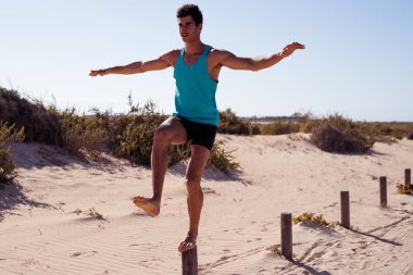 Bir bacakla direğe dengeleyen genç adam ve kolları yana doğru uzatılmış.