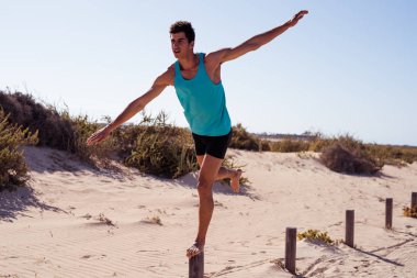 Bir bacağıyla bir direğin üzerinde duran genç adam kollarını yanlara uzatarak öne doğru eğilir.