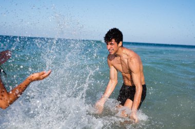 Young man bending in the ocean and splashing a young girl with water in summer