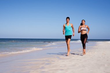 Genç çift sıcak bir yaz gününde kumlu beyaz plajda kıyı boyunca çalışan, her ikisi de egzersiz giysileri giyiyor