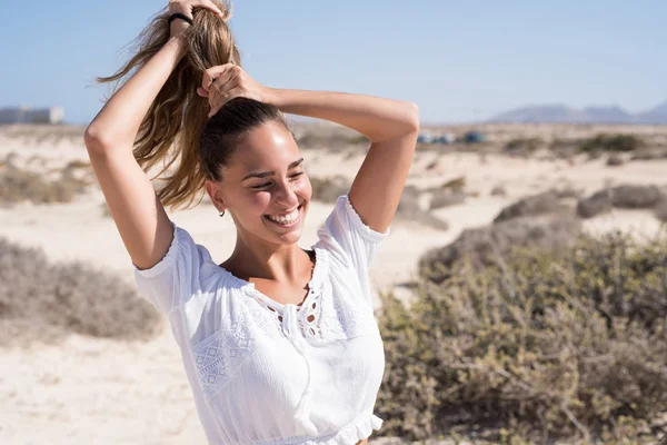 Young woman in a white shirt is standing on the beach and holding her hair above her head in a pony tail