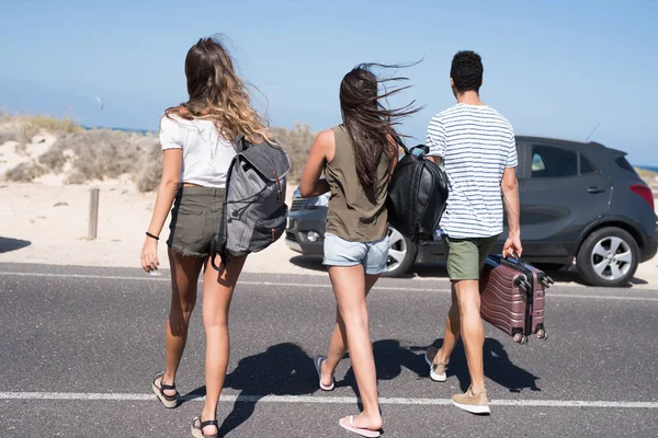 Three young people walking across the road near a beach and next to a parked car , they are all carry bags