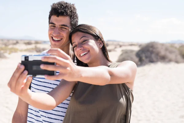 Young millennial woman holding a camera up and taking a photo of herself and her companion as they stand on a sandy white beach