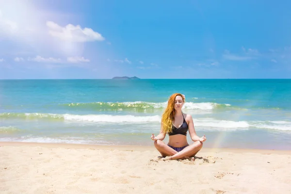 Ein junges, schönes Mädchen mit roten Haaren, im Bikini. praktiziert Yoga auf weißem Sand und türkisfarbenem Meer. — Stockfoto
