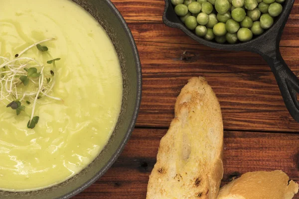 Plano detalle de Sopa de arvejas con pan en mesa de madera rstica y pequeo plato hondo con arvejas en vaina