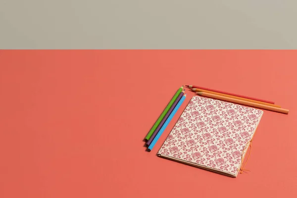 Escritorio en Fondo coral vivo color del 2019 con cuaderno con patron renacentista en rojo burgundy  y beige acompaado de lapices de colores naranja, rosa, rojo, azul, can y verde y pared gris