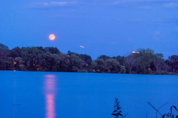 Night time long exposure photo of a full orange moon and reflection over a beautiful lake during summer time. Airplanes light trail streaks across sky on approach to airport