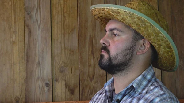 A grown man looks into the distance. Straw hat on his head. He\'s wearing a plaid shirt. Unshaven face.