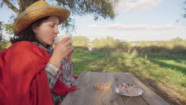 A country woman in a straw hat. Sitting and drinking tea. Big white mug. On the background of a large tree. Blue sky. Green grass and leaves. Country life.