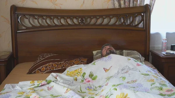 Adult bearded man lies on the bed. It is covered with a color sheet. The interior of the bedroom.