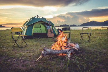 Şenlik ateşi arka planda yeşil alan çayır, göl ve dağ ile kamp çadır. Piknik ve seyahat kavramı. Doğa teması.
