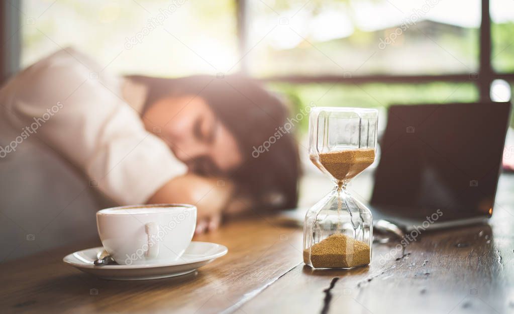 Close up of sandglass with business woman tired from working with laptop computer background. Relax Sleeping and taking nap concept. Business people and overtime workload theme. Office and home theme