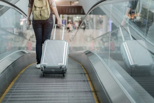 Close up lower body of woman traveler with luggage suitcase going to around the world by plane. Female tourist on automatic escalator in airport terminal.