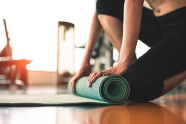 Spor salonunun arka planında yoga şiltesini katlayan sportif bir kadın. Yoga dersinden sonra paspas yuvarlama egzersizi. Spor ve spor eğitimi konsepti. Ellerini halıya koy.