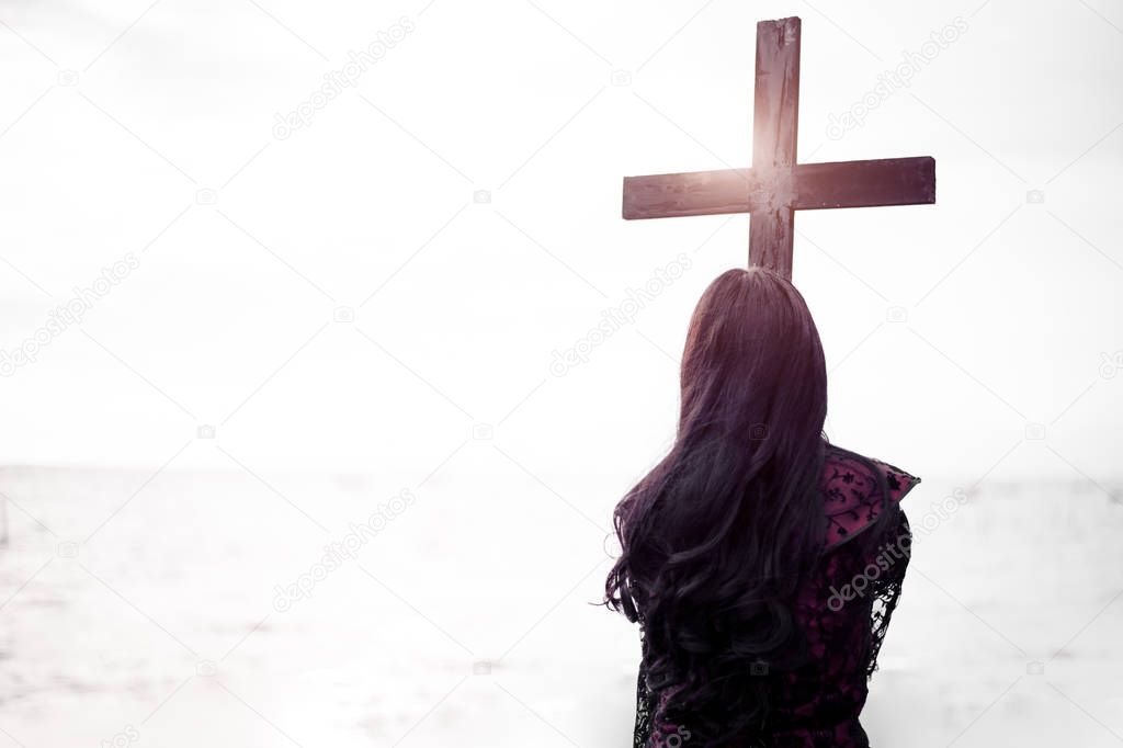 Christian woman in black holding black cross for funeral with sea and sky background. Halloween day and mourning after death of relative people. Dark and Bright tone mixed. Horror and thriller theme.