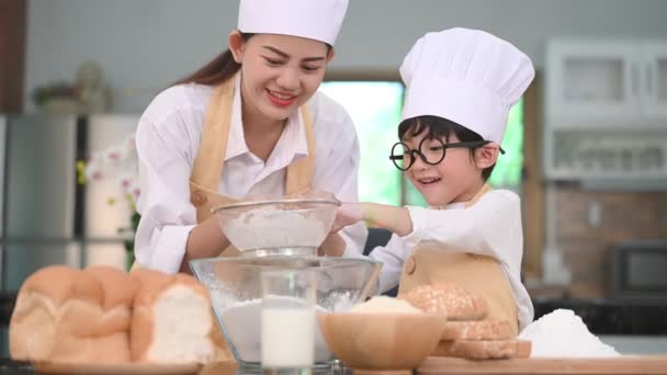 かわいい小さなアジアの男の子と美しい母親は パン屋やケーキを焼く準備のためにテーブルの上の家庭用キッチンでふるいふるいコランダーで生地の小麦粉をふるいにかけます シェフ面白いとして小麦粉で遊ぶタイの子供たち — ストック動画