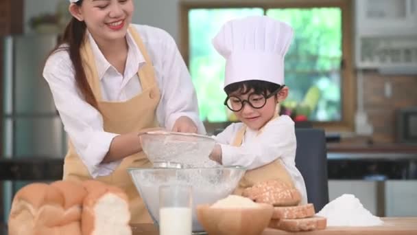 かわいい小さなアジアの男の子と美しい母親は パン屋やケーキを焼く準備のためにテーブルの上の家庭用キッチンでふるいふるいコランダーで生地の小麦粉をふるいにかけます シェフ面白いとして小麦粉で遊ぶタイの子供たち — ストック動画