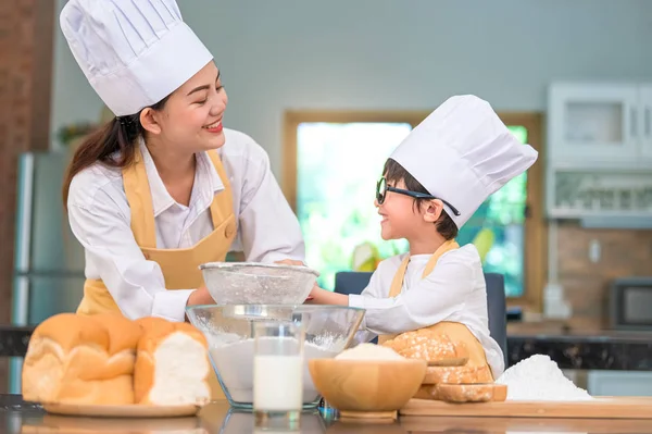 Cute little Asian boy and beautiful mother sifting dough flour w