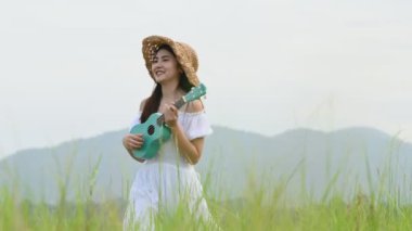Beyaz elbiseli Asyalı mutlu kadın yeşil çayır tarlasında gitar çalıyor ve yaz mevsiminde dağcılık yapıyor. Müzisyen güzel kadın müzik aleti ile rahatla. Kırsal kesimde yaşam tarzı bağbozumu